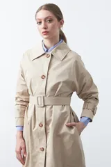Trench de diseño clásico en color beige, con cierre frontal de botones, cinturón ajustable con hebilla a tono y mangas con trabillas en los puños.