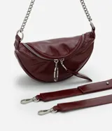 Cartera tipo riñonera de tamaño pequeño, confeccionada en poliuretano con acabado brillante. Presenta un diseño de media luna con cierre de cremallera metálica, decorado con tiradores circulares. Incluye una correa de cadena metálica para llevar al hombro o cruzada.