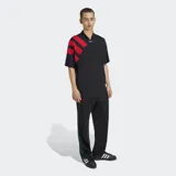 Remera Adidas de estilo futbolístico inspirada en los años 90, color negro con cuello en pico y tres bandas diagonales rojas sobre el hombro derecho. Presenta un corte holgado y logo de la marca en el centro del cuello.