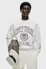 Buzo de algodón con cuello redondo, hombros caídos y puños acanalados. Presenta un estampado frontal con estilo universitario que incluye el texto 'NEW YORK PHYS.ED.' y un emblema central en tonos rojo y azul.
