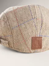 Boina estilo flat cap con diseño de espiga y cuadrillé en tonos beige, azul y rojo. Cuenta con una pequeña hebilla metálica ajustable en el lateral.
