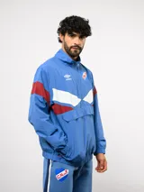 Buzo deportivo de nylon con medio cierre frontal y cuello alto. Presenta un diseño de bloques de color en azul, blanco y bordó, con el escudo del Club Nacional de Football y el logo de Umbro en el pecho.