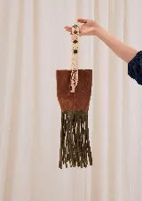 Bolso estilo bucket de cuero marrón con flecos largos verde oliva en la base, correa tejida en macramé crudo con detalles de piedras, broche a presión y costuras visibles.
