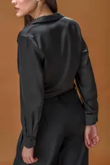 Camisa negra de satén con escote en V cruzado y cierre lateral. Presenta mangas largas con puños abotonados y un diseño fluido que estiliza la silueta.