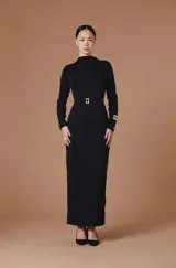 Vestido largo entallado de color negro, con cuello alto y mangas largas. Presenta una textura plisada con ondas verticales y un cinturón integrado con hebilla metálica dorada en la cintura.