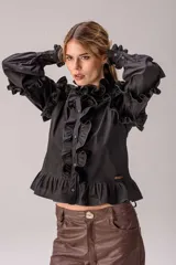 Camisa negra de corte entallado con volados decorativos en el cuello, la botonadura frontal, los hombros y las mangas. Presenta mangas largas con puños ajustados y un ruedo con volado inferior.