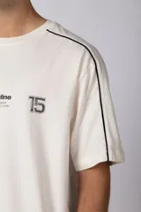 Remera de algodón color blanco con corte regular, cuello redondo y mangas cortas. Presenta detalles de líneas finas negras en los hombros y estampado gráfico frontal con tipografía y logotipos en color negro.