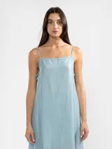 Vestido largo de color celeste confeccionado en tejido arrugado liviano. Presenta un diseño con escote recto, breteles finos y detalles de lazos en los laterales. Incluye bolsillos laterales, forro interno corto y un ruedo asimétrico con caída posterior más larga.