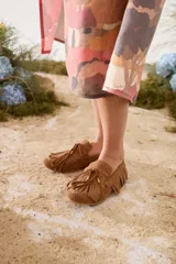 Mocasines de gamuza color tostado, con diseño clásico, flecos alrededor del calzado y en la capellada, y cordón decorativo con detalles metálicos dorados.
