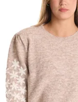 Buzo de punto marca Pinch, color marrón claro, con cuello redondo y mangas largas. Presenta un diseño de flores blancas bordadas a lo largo de las mangas y detalle de frunce en los hombros. Cuenta con puños y ruedo acanalados.