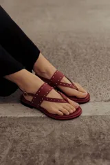 Sandalia plana de cuero color rojo, estilo chinela, con tiras finas adornadas con microtachas doradas. Posee tira de sujeción al tobillo con hebilla.