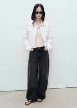 Chaqueta crop estilo denim de color blanco, confeccionada en algodón con elasticidad. Cuenta con cuello camisero, manga larga con puños abotonados, cierre frontal con botones y cuatro bolsillos (dos laterales y dos de parche con solapa en el pecho).