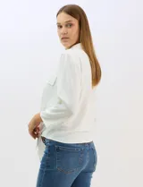 Camisa blanca de rayón con textura sutil, cuello con solapa, cierre frontal con botones y bolsillos simulados en el pecho. Presenta mangas 3/4 regulables con presilla y un diseño con nudo en el ruedo frontal.