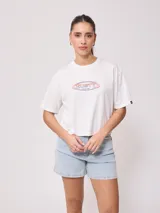 Remera blanca de algodón con calce holgado y largo cropped. Estampado frontal con el logo de la marca Rusty en óvalo con borde rosa y celeste.
