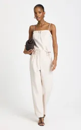 Pantalón sastrero de lino y algodón color beige, de tiro alto y corte ancho (wide leg). Presenta sutiles pliegues frontales, trabillas con apliques metálicos dorados en la cintura y bolsillos laterales.