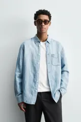 Camisa de corte regular fit confeccionada en denim ligero de algodón con efecto lavado. Presenta cuello solapa con botones, manga larga con puños abotonados, cierre frontal de botones y un bolsillo plastrón en el pecho.