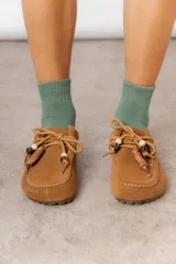 Zueco de cuero gamuzado color tostado, con cordones decorativos adornados con cuentas de madera y piezas envueltas en cuero. Diseño tipo mocasín con costuras visibles en la parte superior y abierto en la parte trasera.