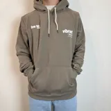 Canguro color caqui con capucha y cordón ajustable. Estampado en la espalda con la frase "The essence of living life fully and energetically", el logo "vibrant journeys", la frase "live for the present" y una cita de Santa Barbara.