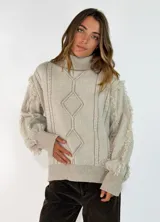 Sweater de cuello alto en color beige, con diseño de tejido trenzado y rombos en el frente. Presenta detalles de flecos en las costuras de las mangas y puños acanalados.