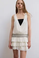 Vestido corto de gasa color blanco, con escote en V cruzado, sin mangas y diseño con volantes en el bajo. Presenta un detalle de tachas plateadas en la cintura.