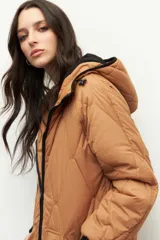 Campera larga color marrón claro, de nylon capitoneado, con capucha, puños en las mangas y forro de polar. Tiene tres bolsillos externos y uno interno.