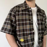 Camisa de manga corta de corte boxy fit, con estampa a cuadros en tonos marrones, azules y blancos. Cuenta con cierre frontal de botones y un bolsillo ojal en el pecho.