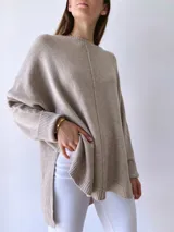 Buzo de tejido de punto color visón, con diseño holgado, cuello redondo, mangas largas con puños acanalados y costura central vertical visible. Presenta un ruedo asimétrico con aberturas laterales.