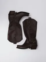 Bota estilo tejana de caña alta, confeccionada en cuero marrón con acabado texturizado. Presenta punta cuadrada, taco bajo y una correa con hebilla metálica ajustable y estribo desmontable en la zona del tobillo.