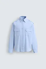 Camisa de algodón color celeste, corte relaxed fit, con cuello solapa, manga larga y trabillas en los hombros. Presenta dos bolsillos de plastrón con solapa en el pecho.