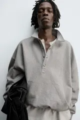 Sudadera de corte boxy fit en color gris, confeccionada en tejido de algodón con interior afelpado. Presenta cuello solapa, cierre frontal con botones a presión, manga larga con puños acanalados y bolsillo frontal tipo canguro. El bajo cuenta con un acabado elástico.