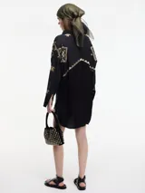 Camisa negra de manga larga con cuello camisero y bordados geométricos en hilo dorado. Presenta un borde en zigzag dorado en la abertura frontal y un largo irregular en la espalda.