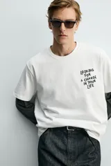 Camiseta blanca de corte relajado, con cuello redondo y manga corta. Presenta un pequeño estampado de texto en contraste en la parte frontal izquierda del pecho.