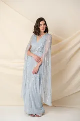 Vestido largo de tul en color celeste pastel, con escote en V y breteles anchos. Presenta un diseño bordado con lentejuelas y pedrería en líneas verticales y una capa larga integrada que nace desde los hombros.