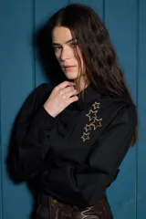 Camisa de corte clásico color blanco, con mangas largas abullonadas en los hombros y puños abotonados. Presenta un detalle de estrellas bordadas en color dorado sobre el lado izquierdo del pecho.