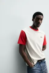 Remera de corte regular fit en color blanco con cuello redondo y mangas cortas en color rojo contrastante, y aplicación de parche bordado en el pecho.