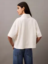 Chomba color crema de punto de algodón con cuello clásico, tapeta de botones oculta y mangas cortas. Presenta un corte holgado y un escudo bordado con el logo de Calvin Klein en el pecho.