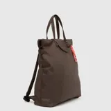 Mochila convertible en tote bag de diseño minimalista, color verde oscuro con acabado impermeable. Cuenta con asas superiores y detalles en cuero sintético gris, cierre superior y un llavero decorativo con la letra B en color turquesa.