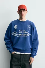 Sudadera de corte boxy fit en color azul con efecto lavado, cuello redondo y manga larga. Presenta estampado frontal con tipografía variada y gráficos estilo universitario. Interior afelpado y acabados en rib.