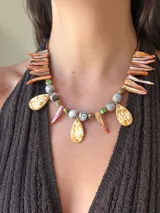 Collar corto de diseño artesanal compuesto por colmillos de nácar teñidos en tonos anaranjados, gotas de cristal checo con efecto jaspeado y cuentas facetadas en tonos verdes y tierra. Presenta cierre de mosquetón y cadena de alargue en acero quirúrgico.