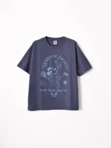 Remera de manga larga con diseño superpuesto que simula una camisa de rayas debajo. La prenda principal es de color azul piedra con estampado gráfico de Minnie Mouse y texto alusivo al tenis, mientras que las mangas y el cuello son de tela camisera a rayas celestes y blancas.