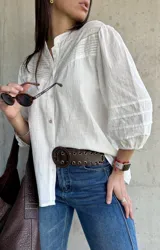 Camisa blanca de estilo romántico con cuello mao, botones frontales y mangas tres cuartos abullonadas con detalle de frunces. Presenta un diseño con textura sutil y costuras decorativas en la parte superior delantera.
