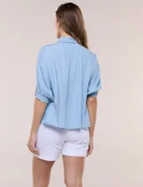 Camisa de tencel liviano estilo denim, color celeste, con cuello camisero, cierre frontal con botones nacarados, mangas cortas abullonadas con puños elásticos en punto smock y detalles de ojalillos.
