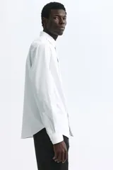 Camisa de vestir blanca con cuello inglés, tapeta clásica y corte ajustado. Presenta mangas largas con puños abotonados y bajo levemente redondeado.