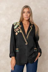 Blazer negro de corte recto con solapas decoradas con ribete dorado trenzado. Presenta bordados artesanales coloridos en el hombro derecho y detalles de flecos con cuentas turquesas en los bolsillos frontales. Cuenta con cierre de un solo botón.