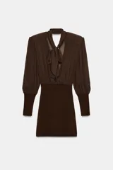Vestido corto de color marrón con diseño combinado. Presenta un cuerpo superior fluido con cuello subido, lazada frontal ajustable y mangas largas abullonadas con hombreras y puños ajustados. La parte inferior es entallada con tejido elástico a tono, espalda con escote fluido y cierre lateral con cremallera oculta.