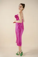 Vestido midi fucsia de satén, con escote buche, breteles finos y detalle de rosa de tela aplicada en el escote. Presenta un tajo lateral en la falda y calce al cuerpo.