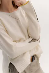 Buzo de lambswool color beige, con escote redondo y detalle en V. Presenta cierres metálicos en los puños que revelan un interior en contraste de color marrón y ribete rojo. Tiene tajos laterales para mayor comodidad y un fit amplio.