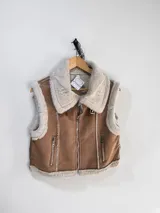 Chaleco estilo gamulán con interior y bordes de piel sintética. Presenta cierre frontal metálico, bolsillos laterales con cierre y detalles de costuras visibles.