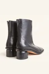 Bota de caña media confeccionada en cuero vacuno negro, con diseño minimalista y cierre trasero. Presenta un taco cilíndrico de madera tallada y punta cuadrada.