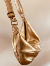 Cartera bandolera tipo banana bag color dorado, confeccionada en cuero blando. Tiene correa ajustable para usar al hombro o cruzada.